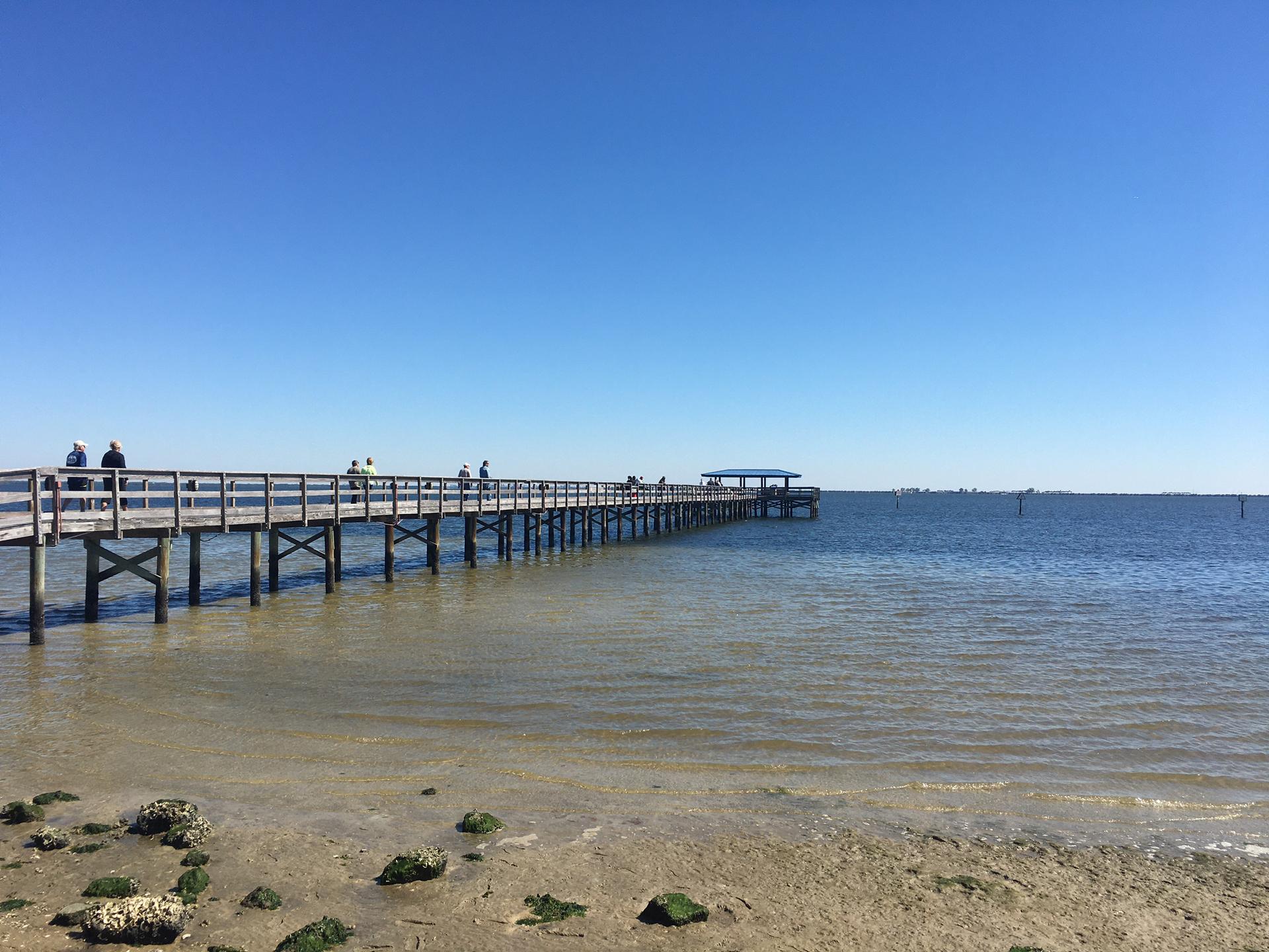 People walk on a long wooden pier over shallow water under a clear sky.