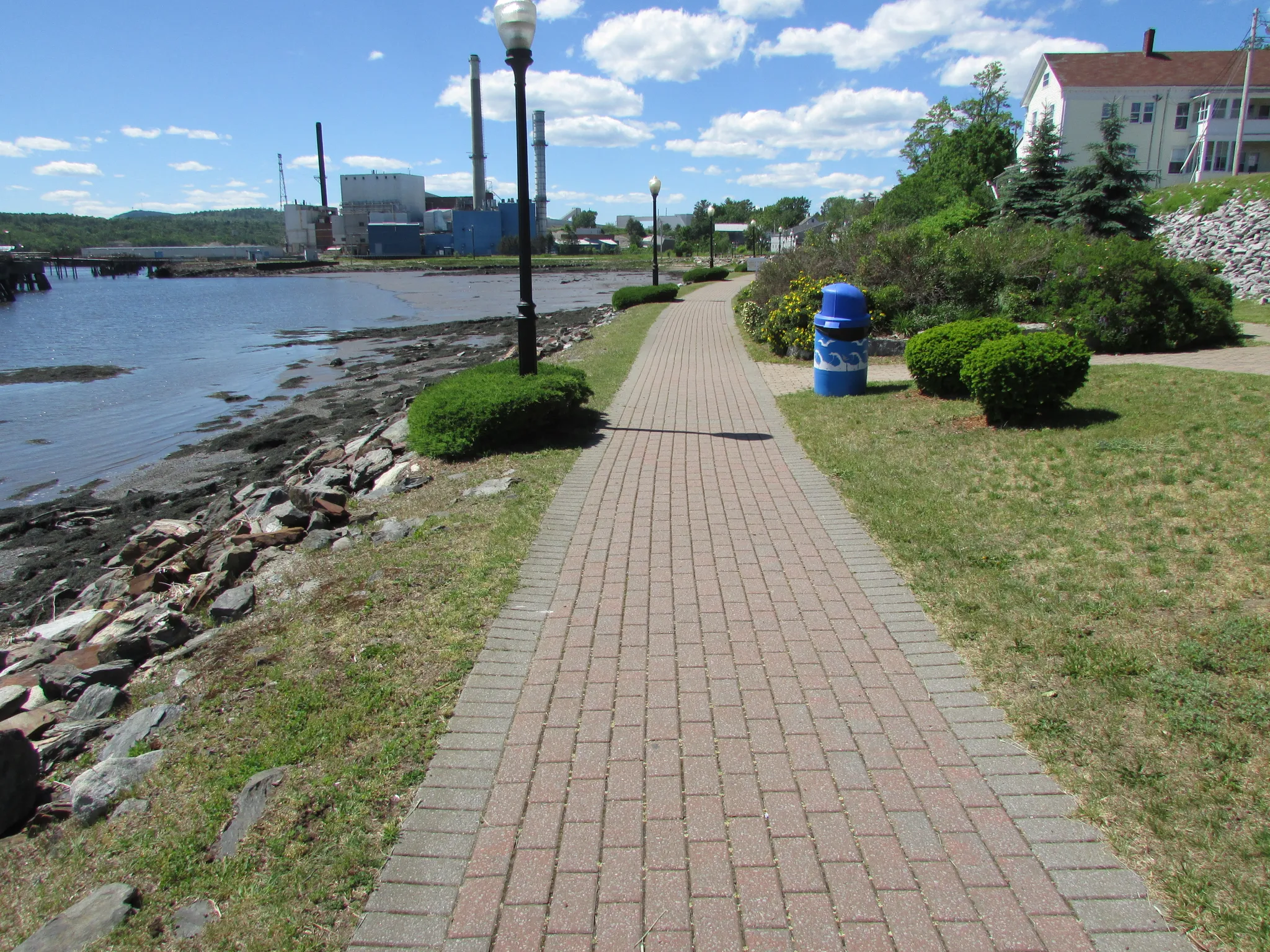 Brick pathway along a sunny waterfront with industrial buildings and green landscaping. - Bucksport Waterfront Walkway