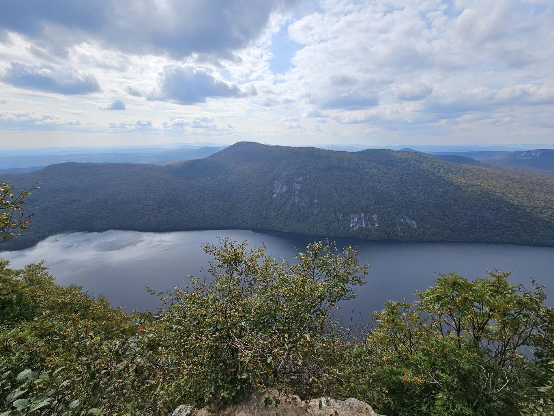 Expansive view of a large lake surrounded by forested mountains under cloudy skies.