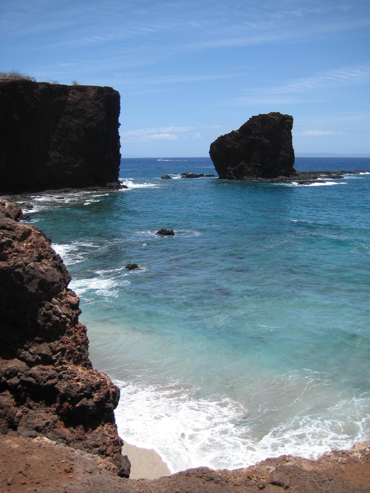 Dark rocky cliffs and sea stacks overlook clear blue ocean on a sunny day.