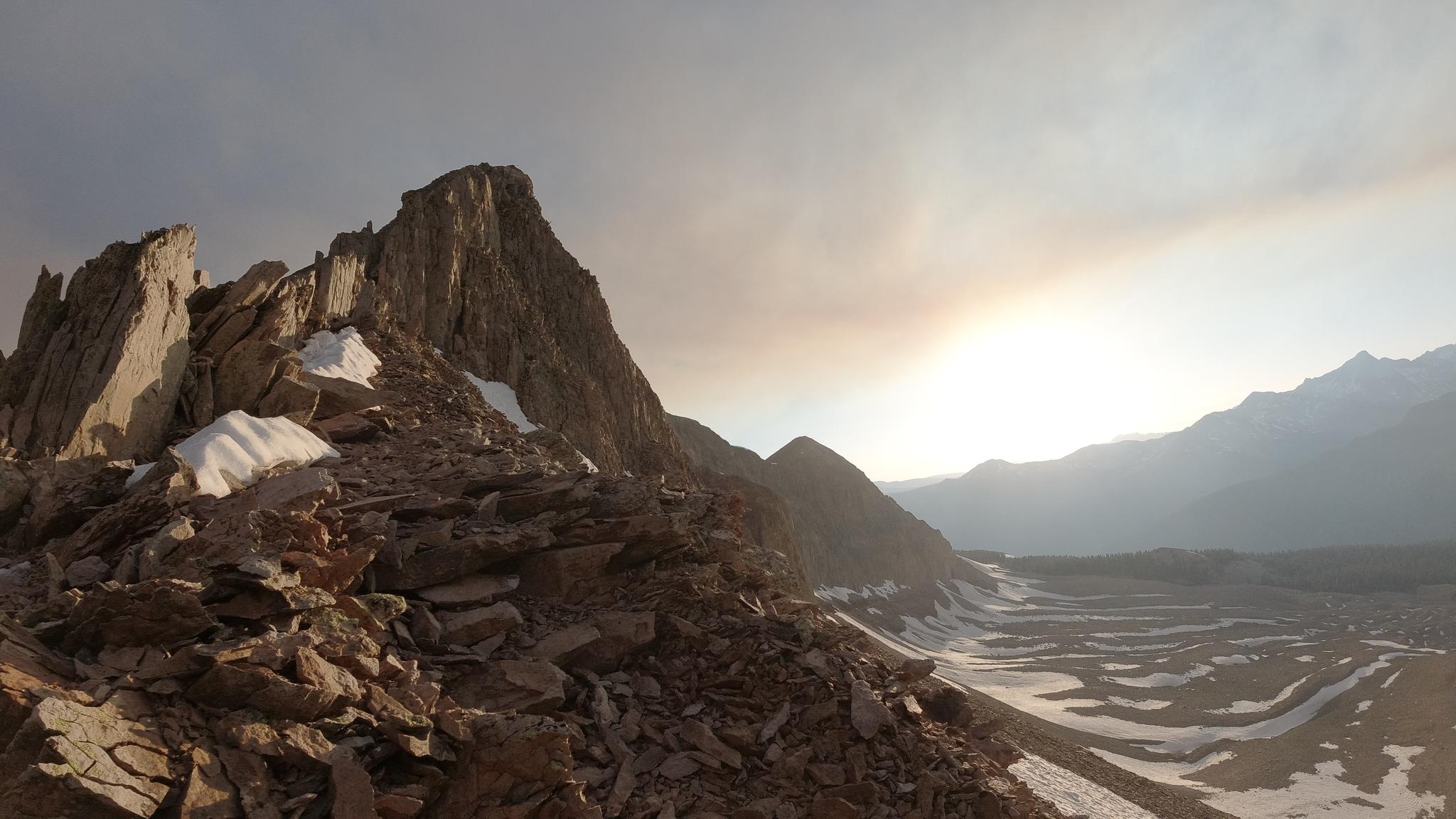 Rugged rocky mountain peak with snow, overlooking a vast valley at sunset.