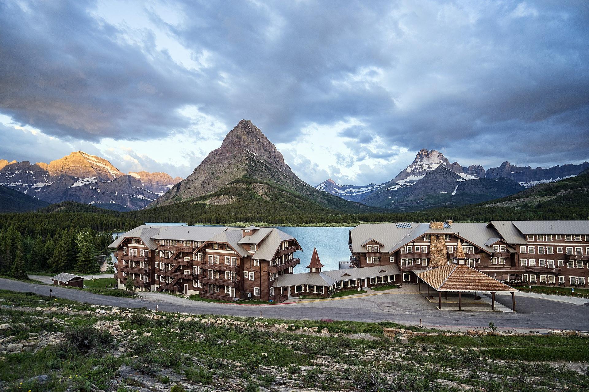 Large lodges sit by a lake with majestic mountains under a cloudy sky.