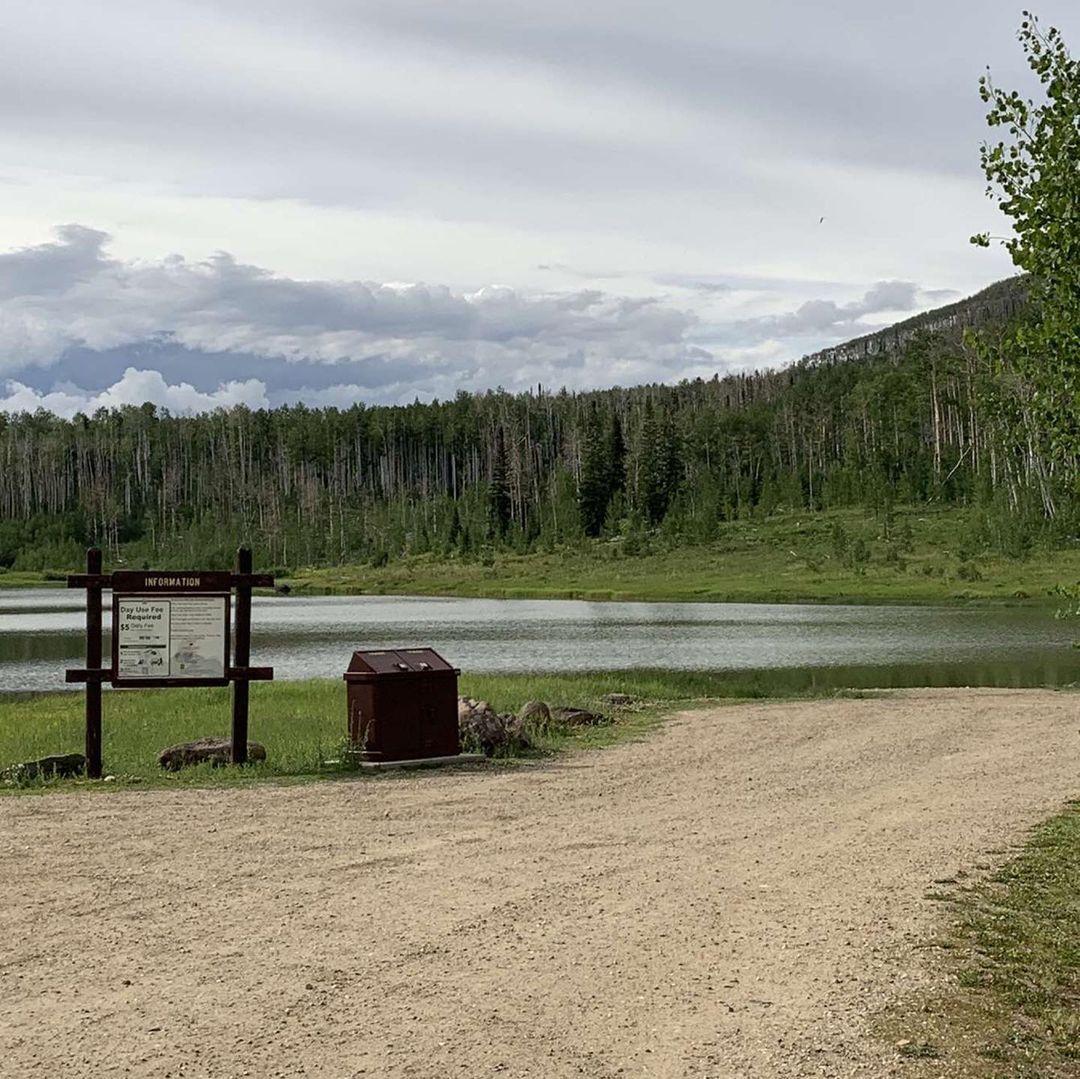 Overcast view of a lake, dirt path, and forested mountains with an information sign.