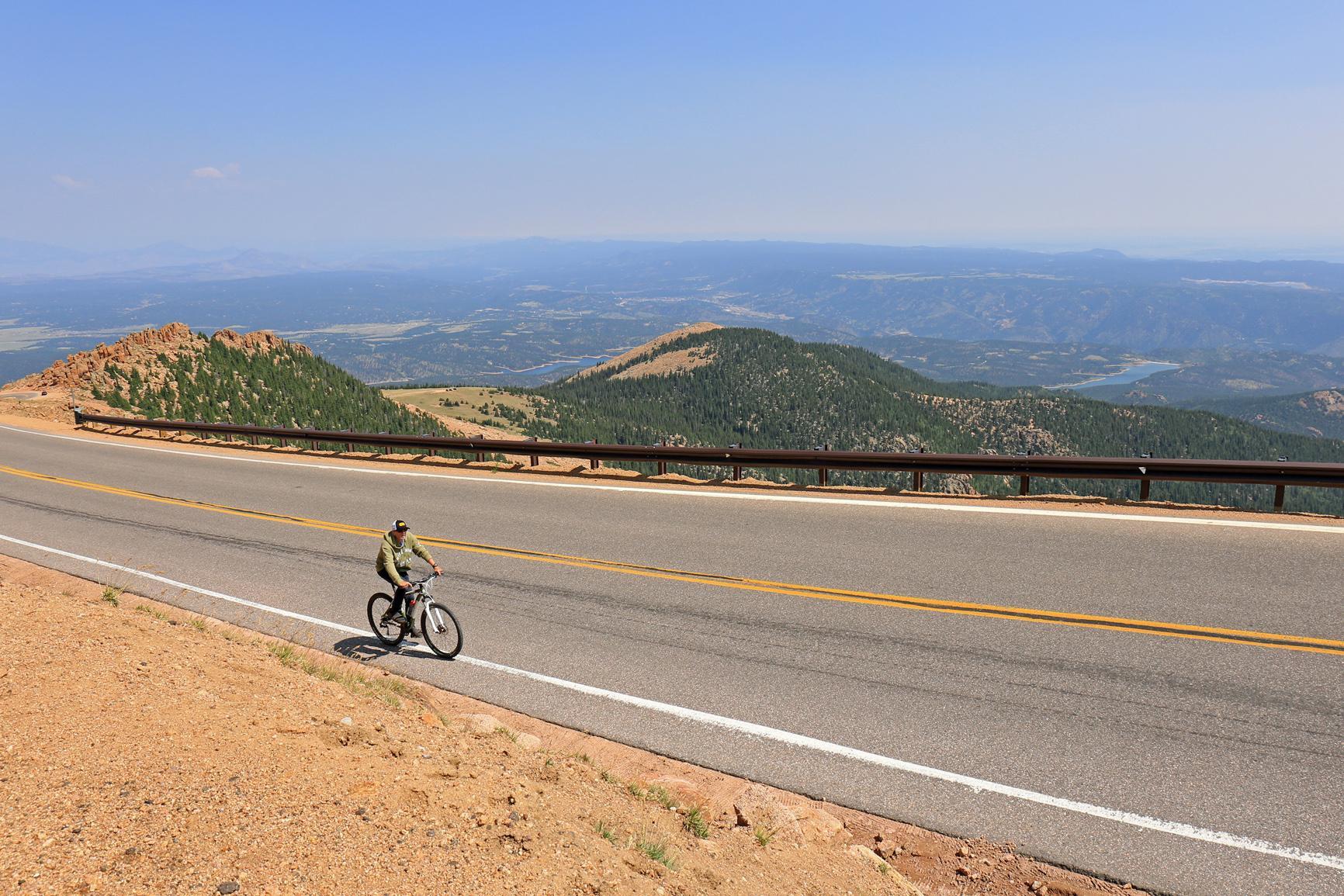 Cyclist on a mountain road with distant forested hills under clear sky.
