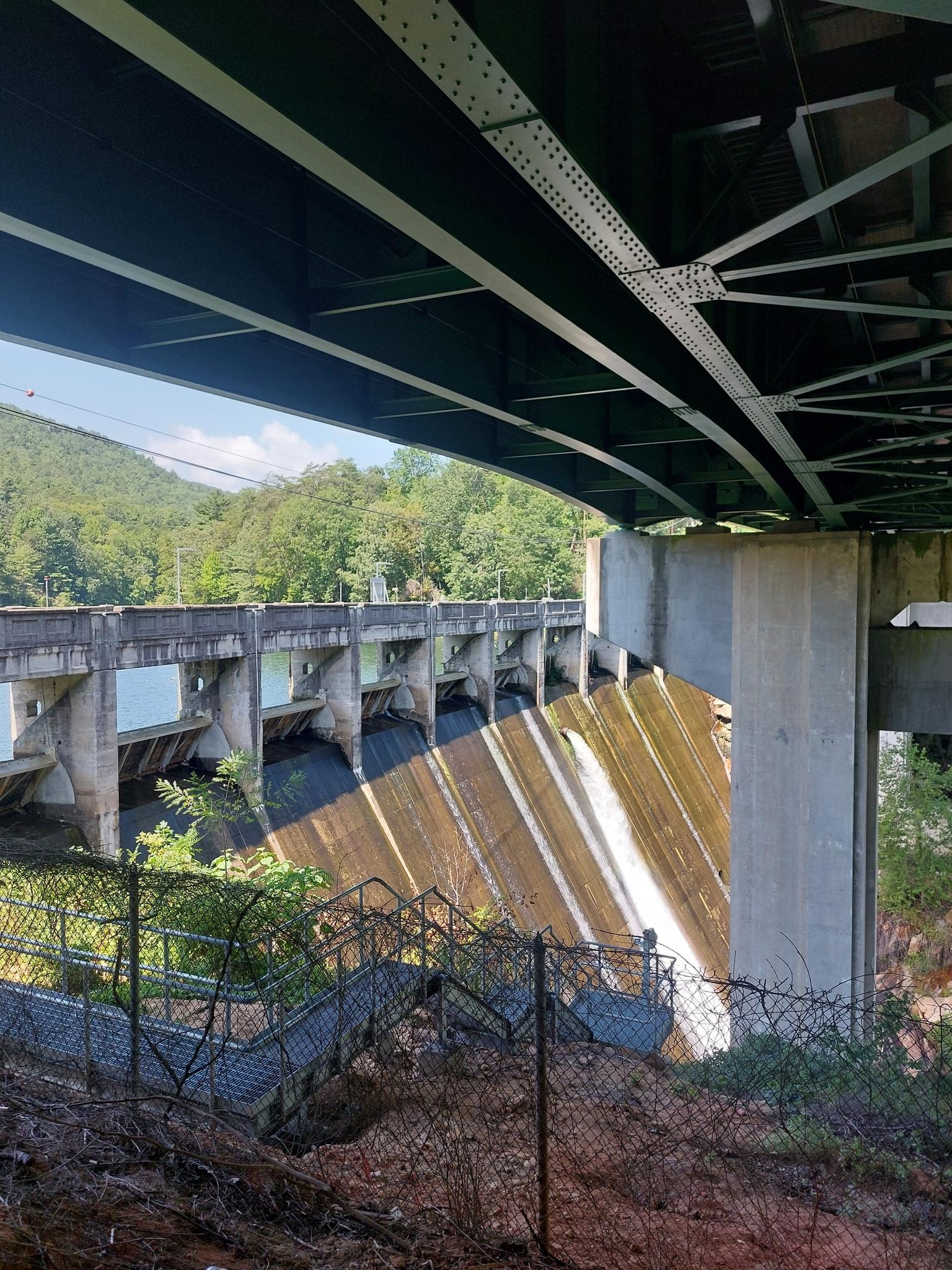 Concrete dam with flowing water, viewed from under a large bridge structure.