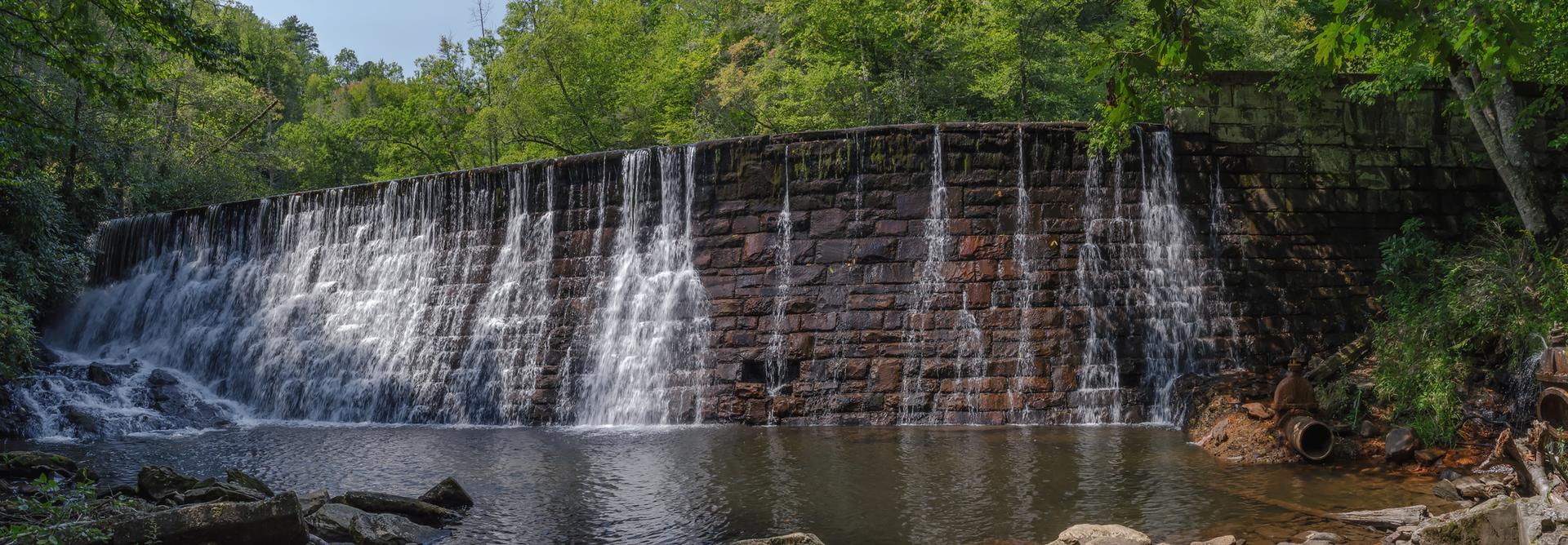Daylight illuminates a stone dam with water flowing over it into a calm pond.