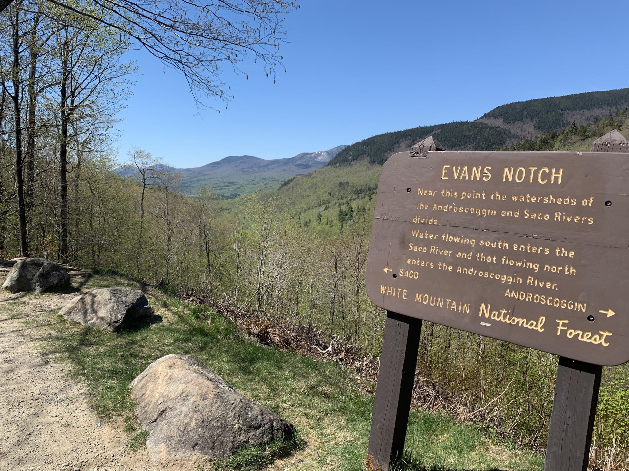Wooden sign on a dirt path overlooking a vast green mountain valley.