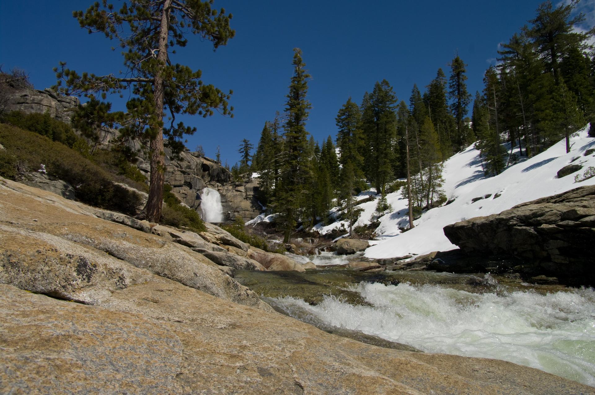 A rushing river flows past snowy banks and pine trees under a clear blue sky.