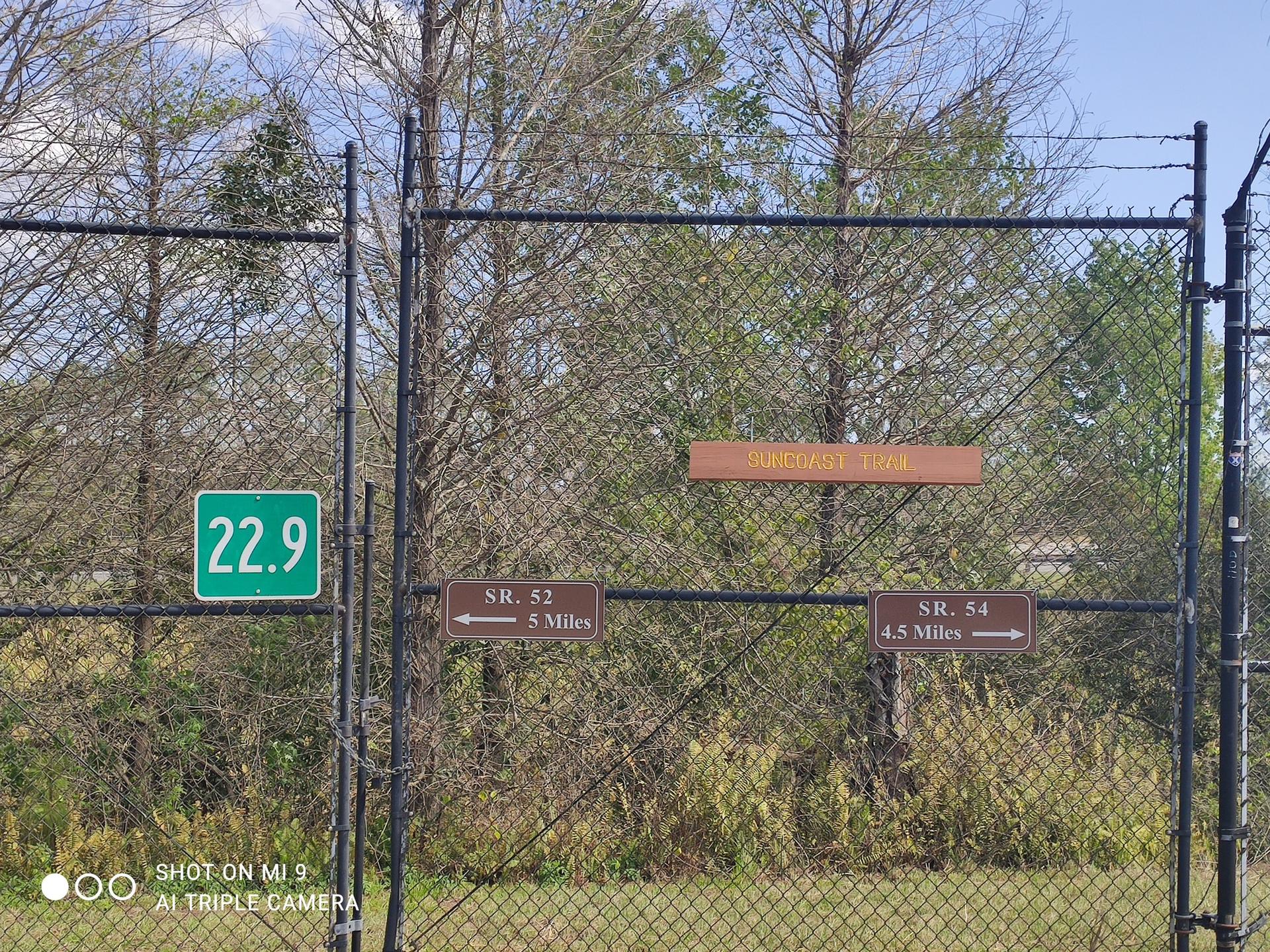 A chain link fence with several directional signs and a green marker in daylight.