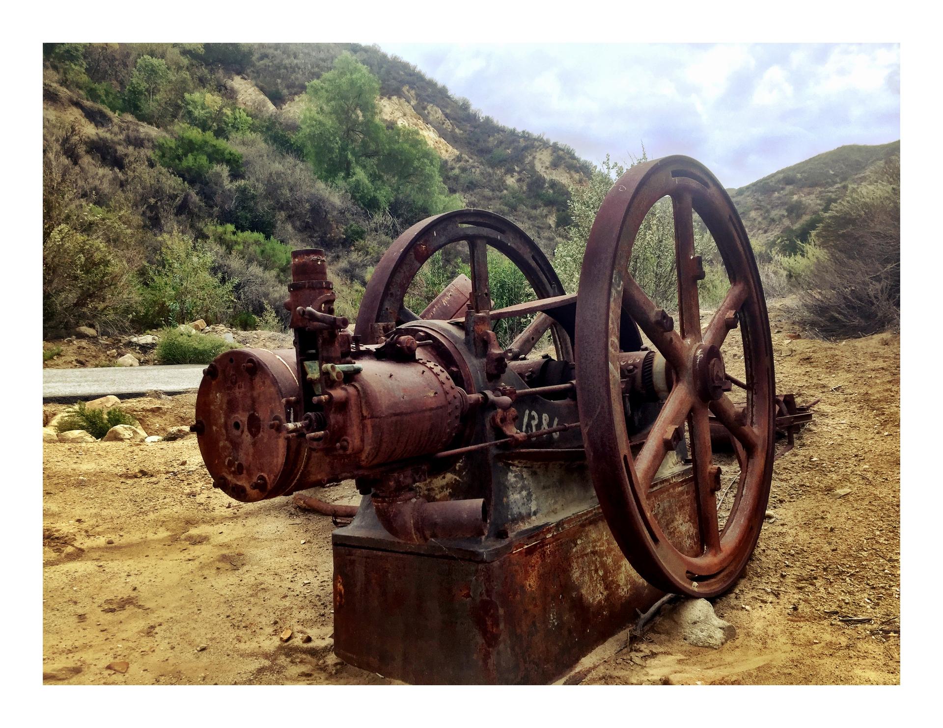 Rusty old machinery with large wheels on dirt ground, hills in background.
