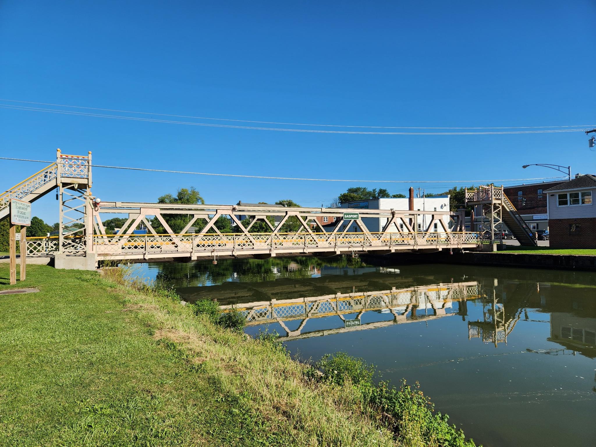 Ornate metal bridge spans calm water, reflecting under a clear blue sky.