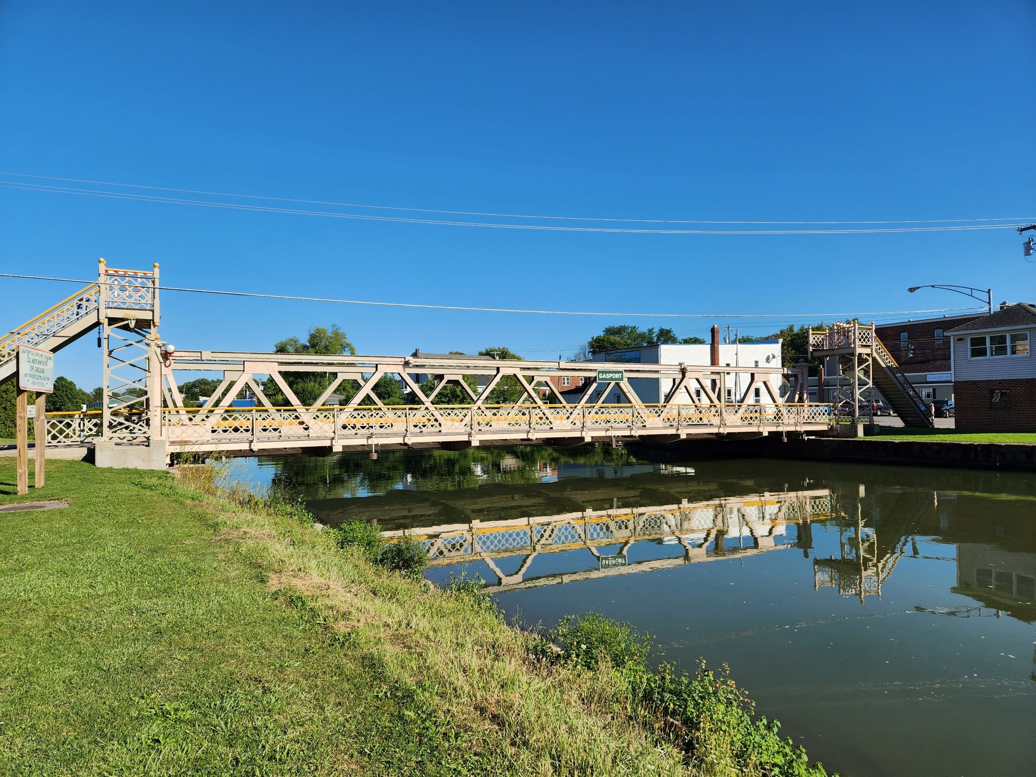 Ornate metal bridge spans calm water, reflecting under a clear blue sky. - Erie Canalway Trail