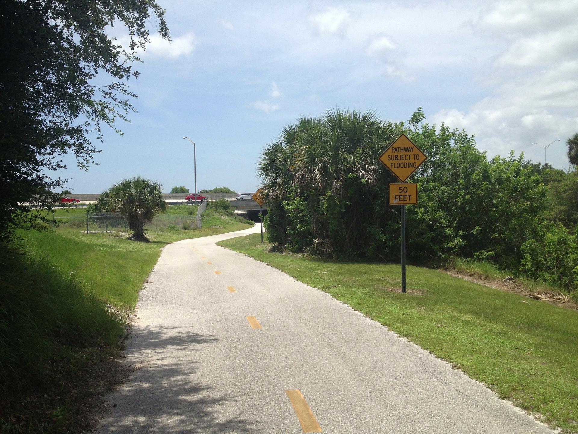 A sunny paved path curves right, bordered by green grass and trees.