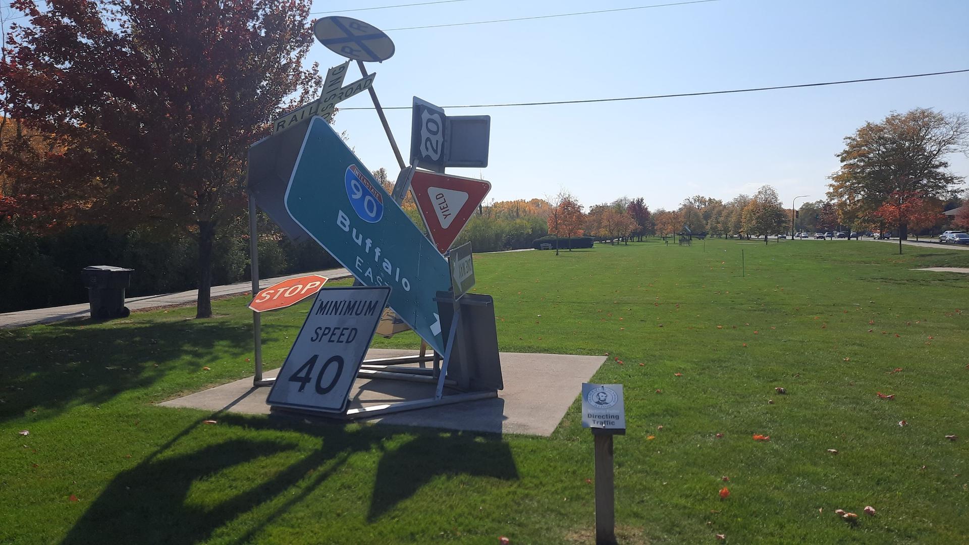 A sculpture made of road signs stands in a sunny, grassy park.