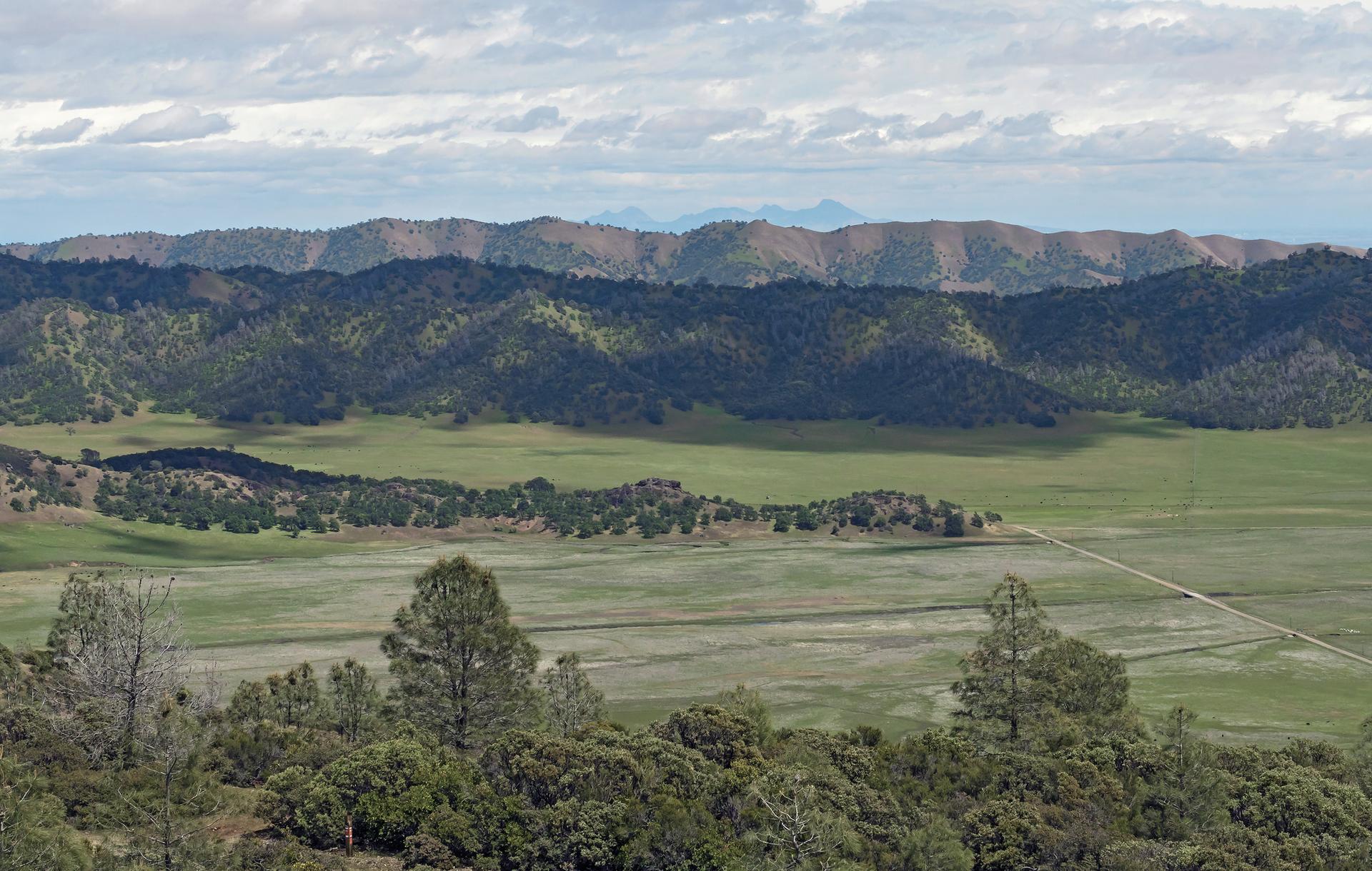 Expansive landscape with green fields, tree-covered hills, and distant mountains under a cloudy sky.