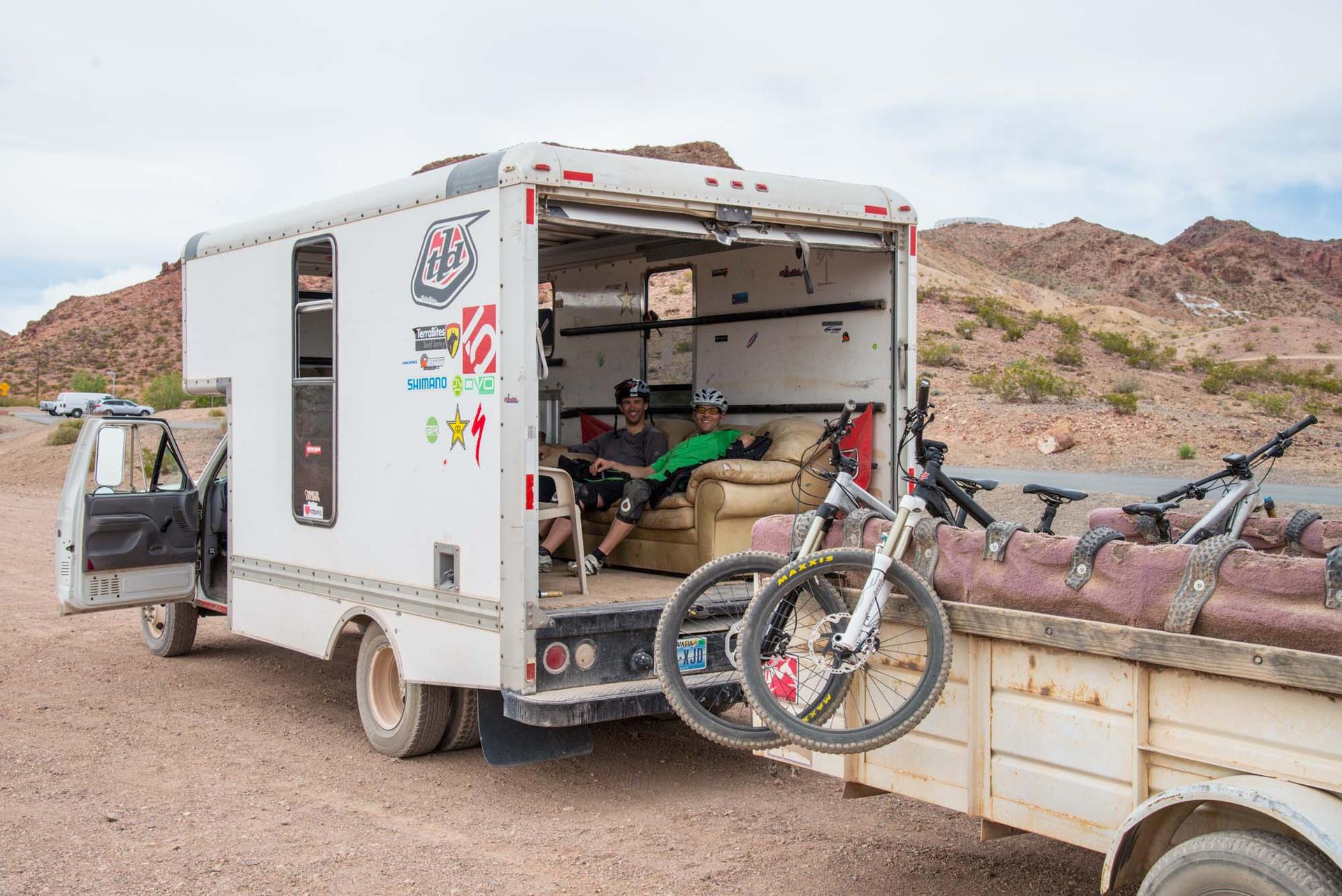 Two people relax in a truck's open back, mountain bikes on a trailer.