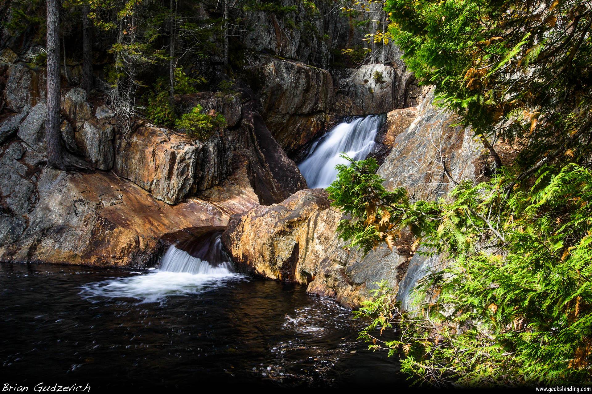Bright waterfall flowing over dark rocks into a dark pool, surrounded by green trees.