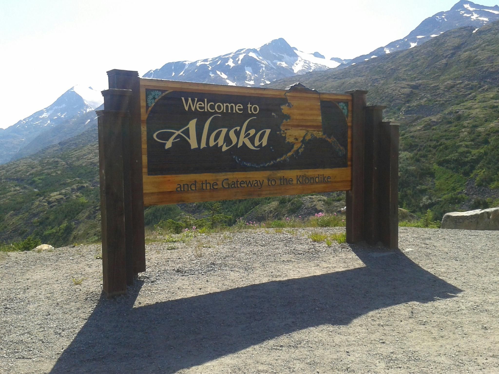 Large wooden welcome sign stands before sunlit, snow-capped mountains. - Skagway to White Pass