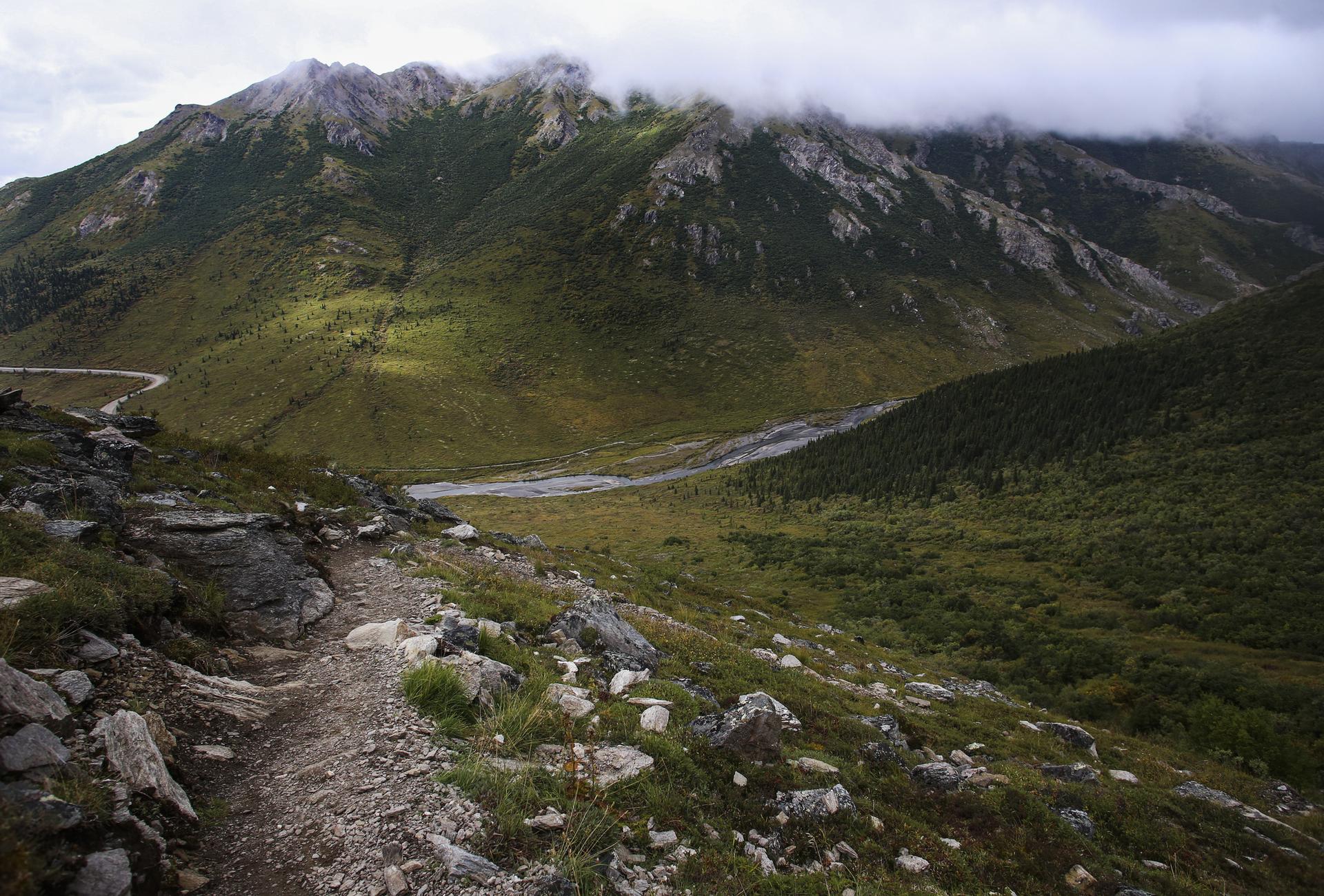 Rocky trail descends into a green valley with a river and cloudy mountains.