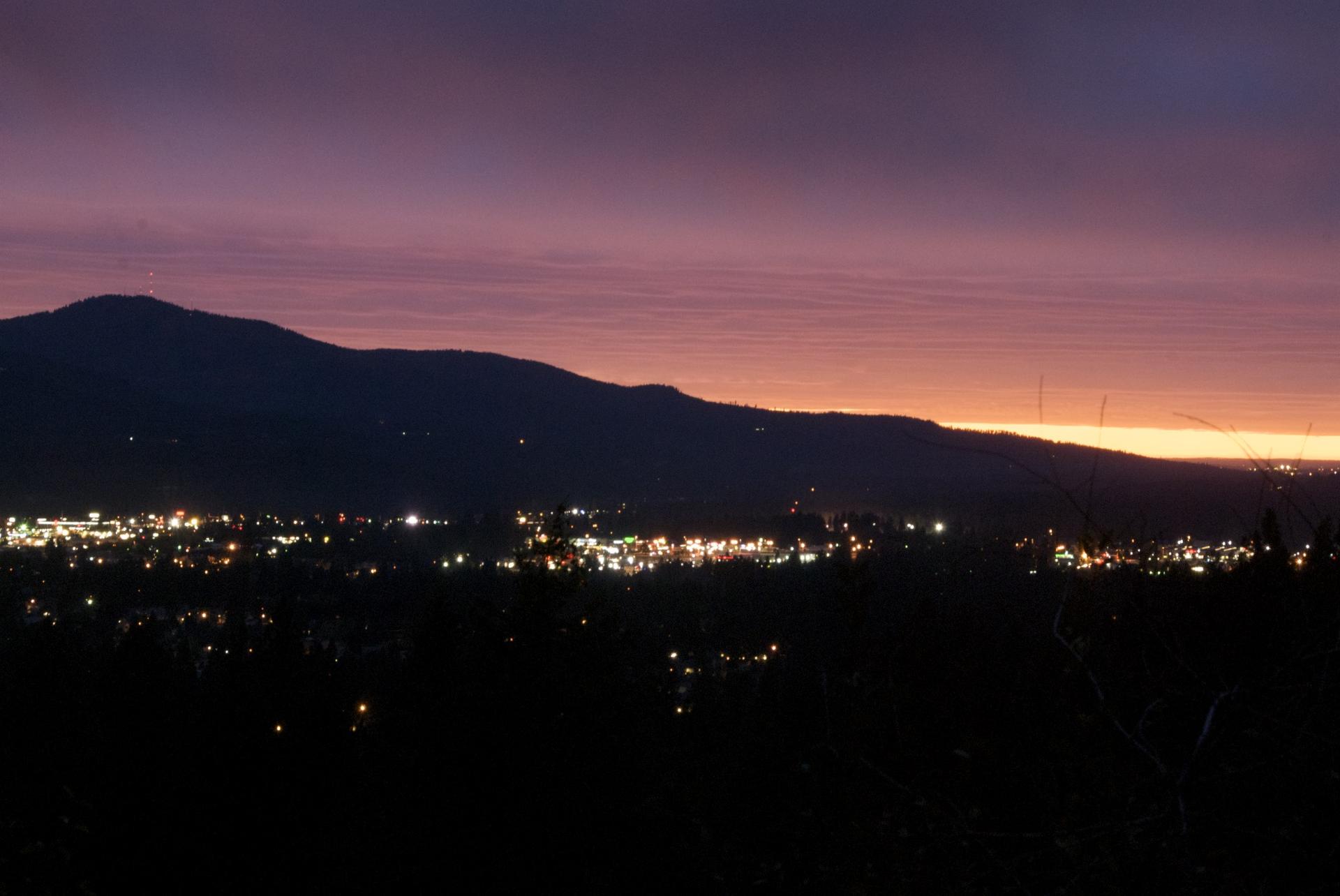 City lights glow below dark mountains under a vibrant purple and orange sunset.