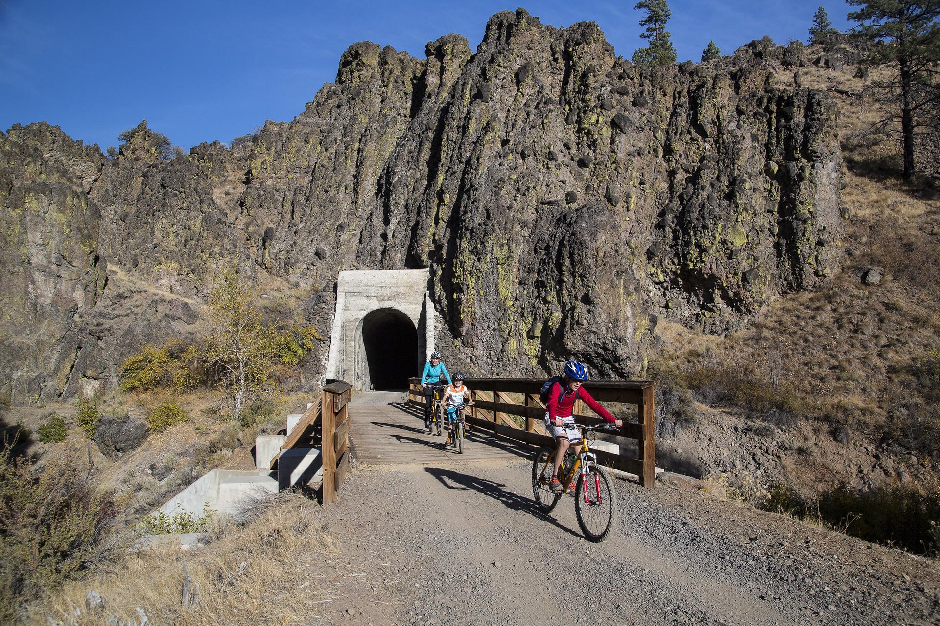 Three cyclists approaching a tunnel entrance carved into a rocky cliff.