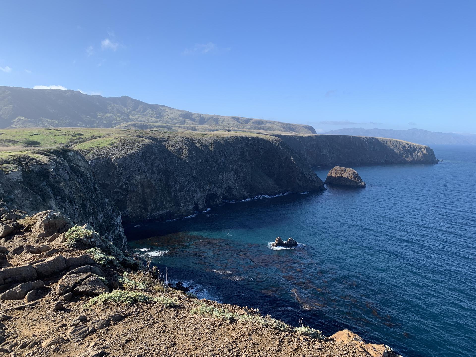 Rugged coastline with dark cliffs, blue ocean, and distant mountains under a clear sky.