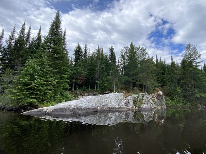 Rocky shoreline with evergreen trees reflected in calm water under a cloudy sky.
