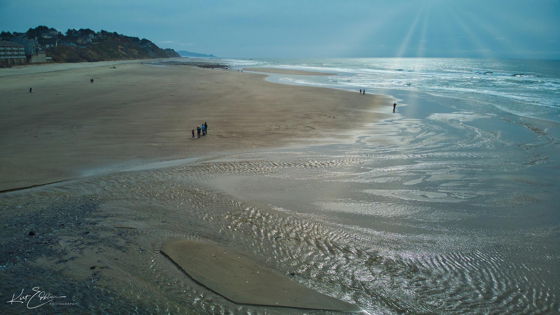 A wide sandy beach with ocean waves, people, and sun rays.