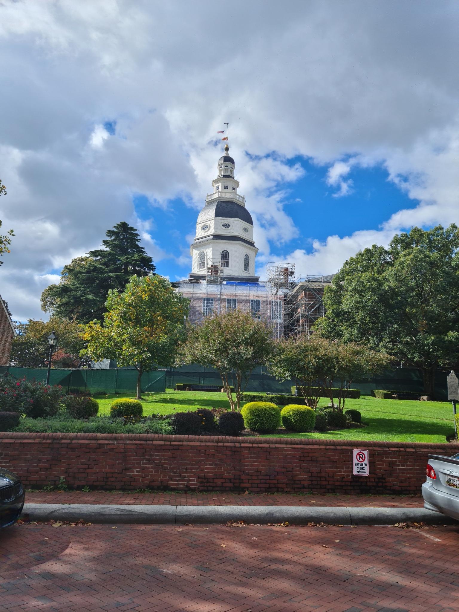Grand domed building with scaffolding, surrounded by green trees and lawn.