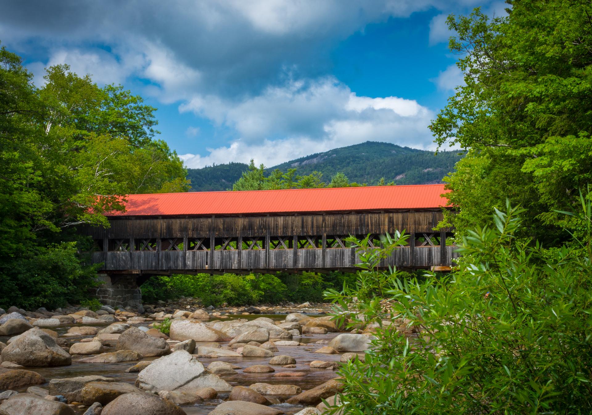 A red-roofed covered bridge spans a rocky river, surrounded by green trees.