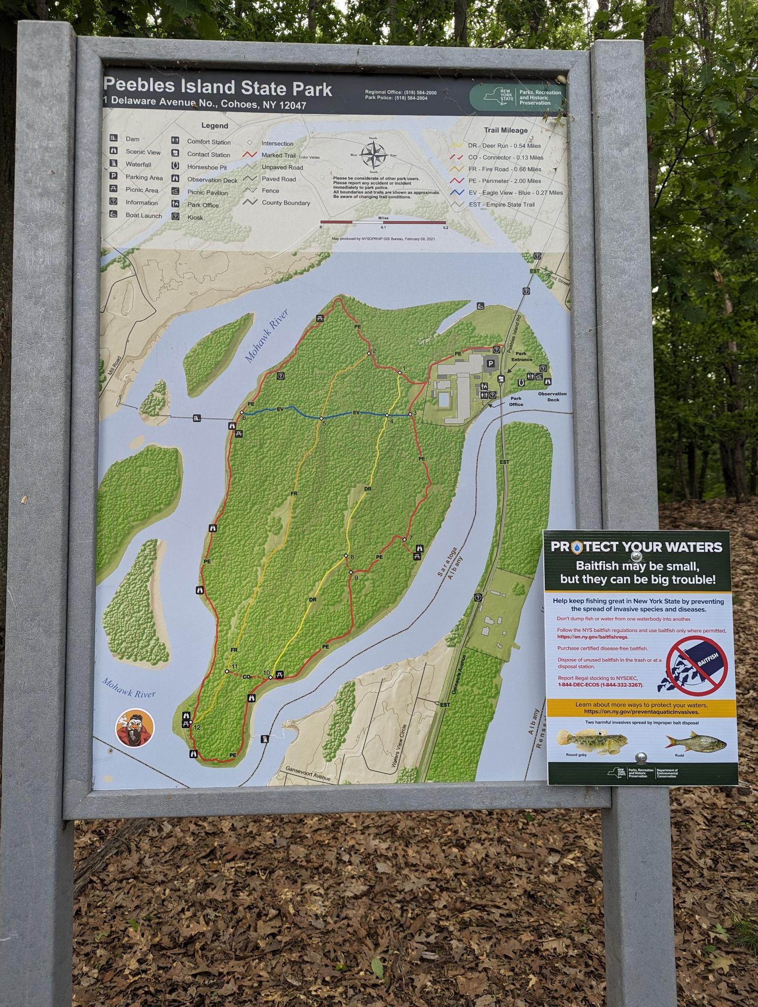 A large park map sign stands on a wooded path with fallen leaves.