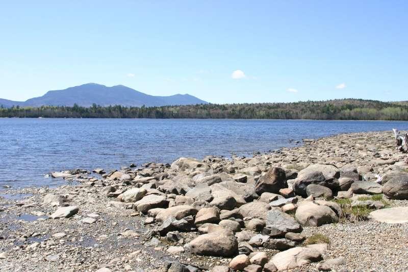 Sunny lake with rocky shore and distant mountains under a clear sky.