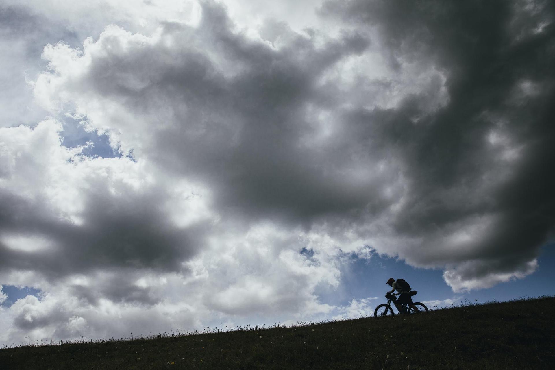 Cyclist silhouetted against a dramatic cloudy sky on a grassy hill.