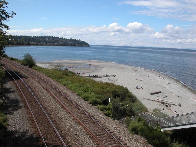 Train tracks run alongside a wide sandy beach with people and calm bay.