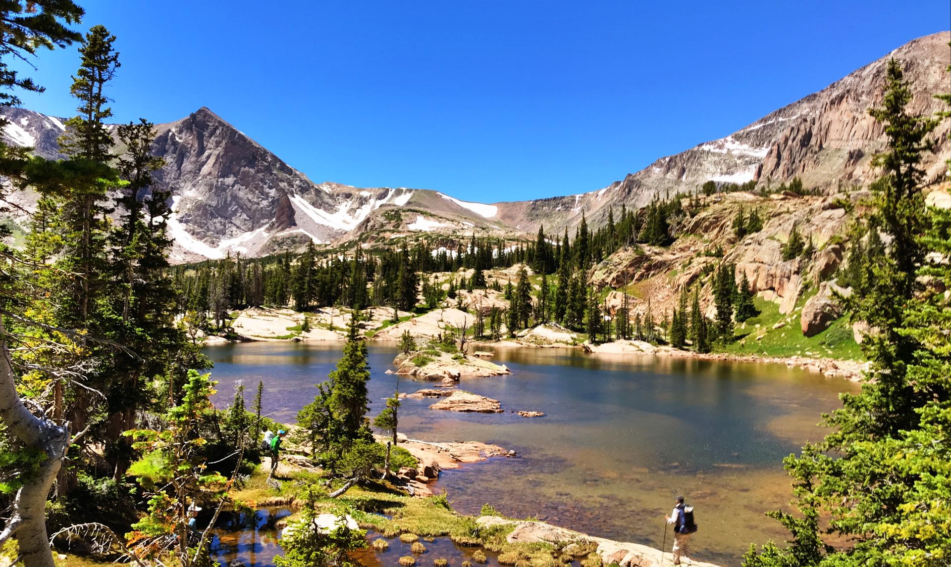 Sunny mountain lake with islands, surrounded by pine trees and snowy peaks.
