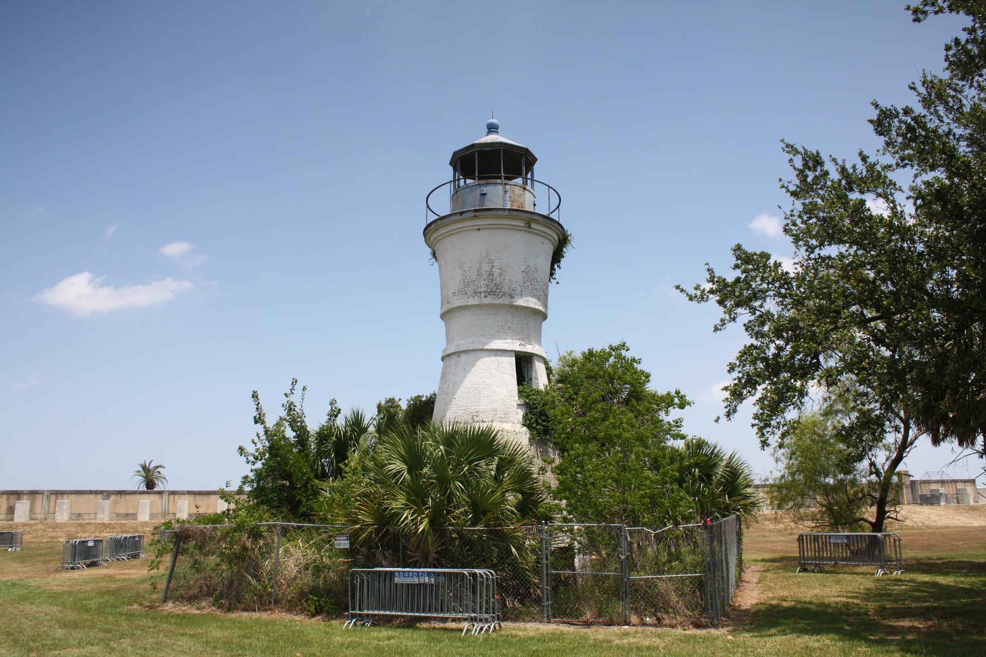 An old white lighthouse stands amidst green bushes under a clear blue sky.