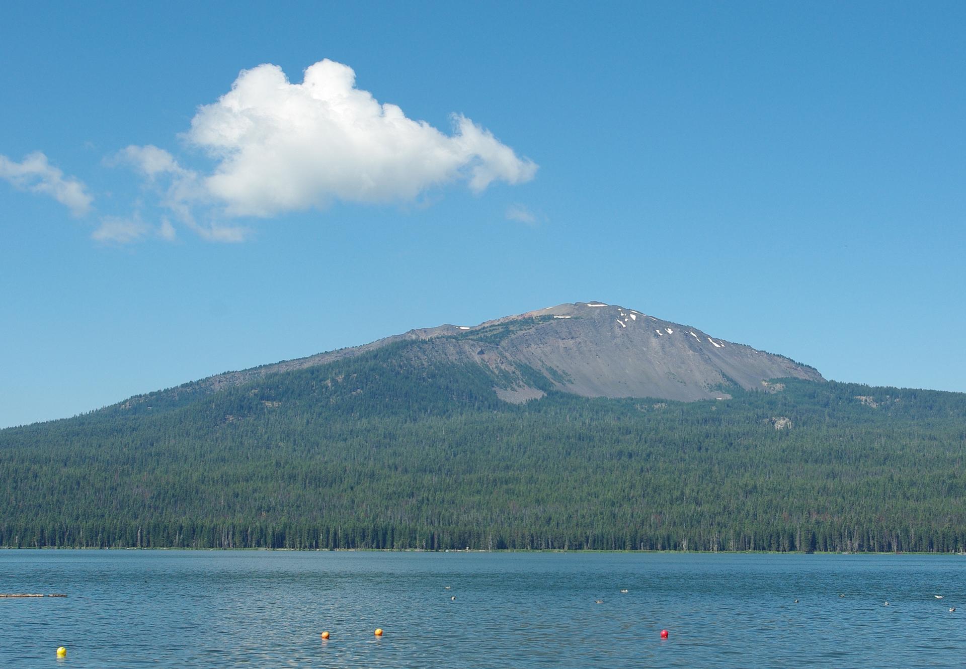 Sunny day with a forested mountain, rocky peak, and calm lake with buoys.