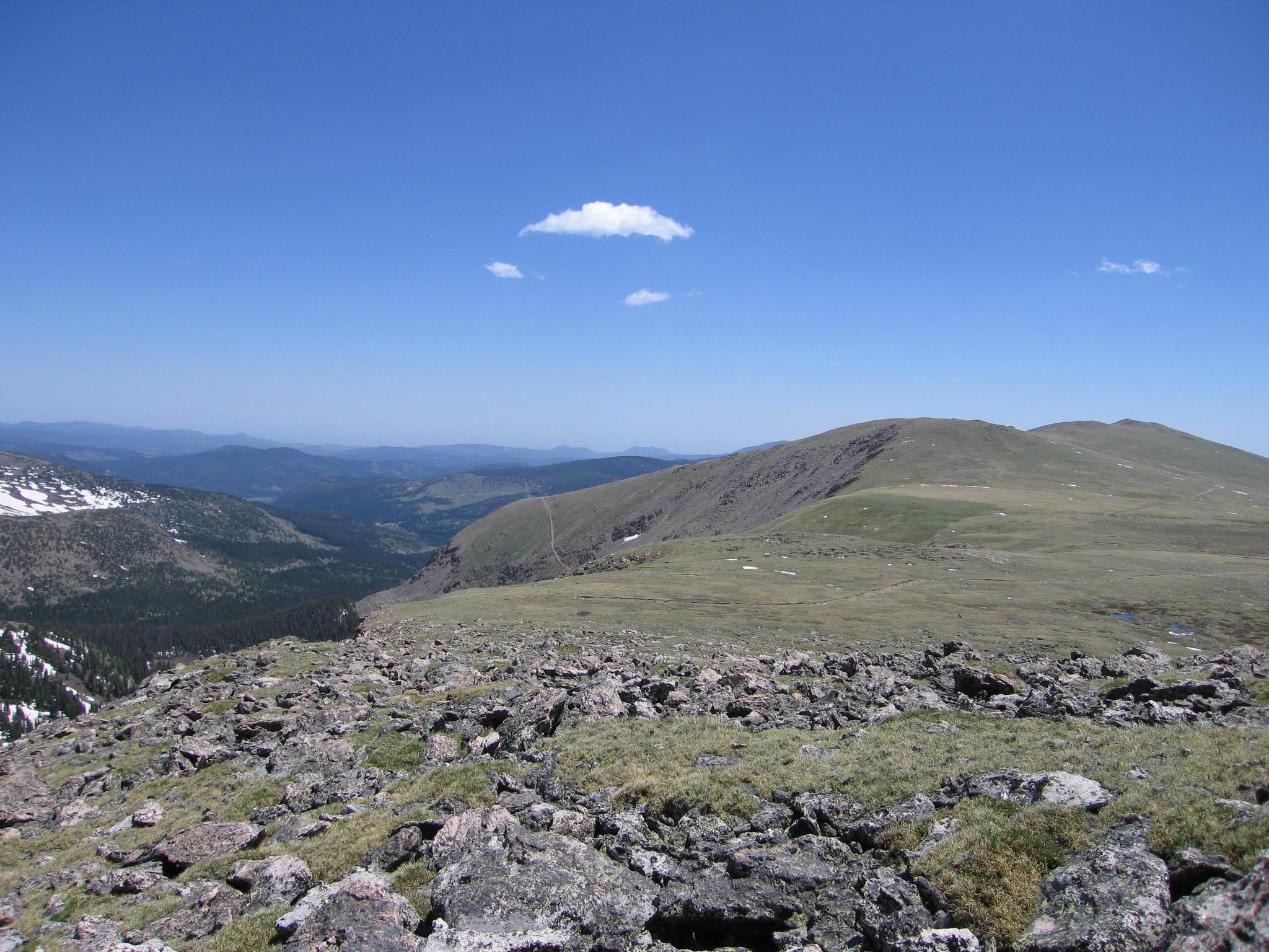 Expansive mountain view with rocky foreground, green slopes, and a bright blue sky.