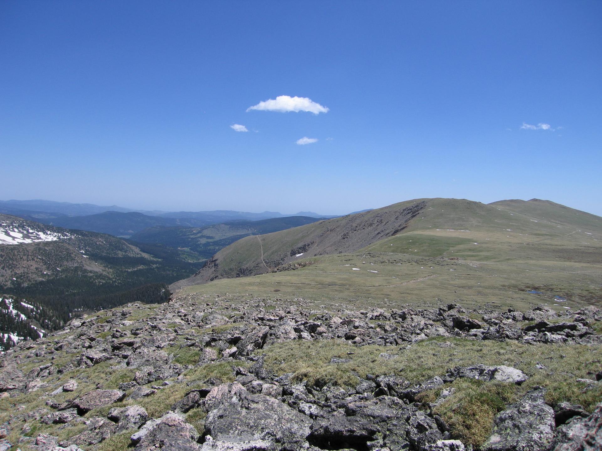 Expansive mountain view with rocky foreground, green slopes, and a bright blue sky.