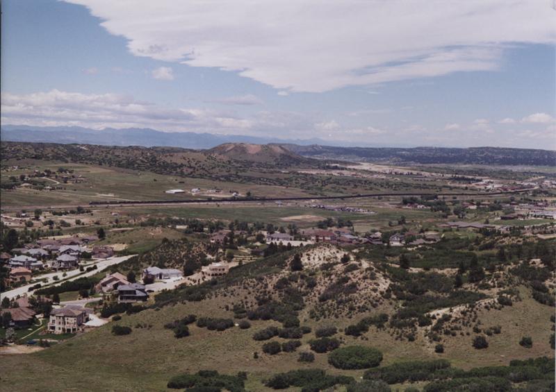 Sprawling suburban landscape with houses, green hills, and a distant train under cloudy skies.