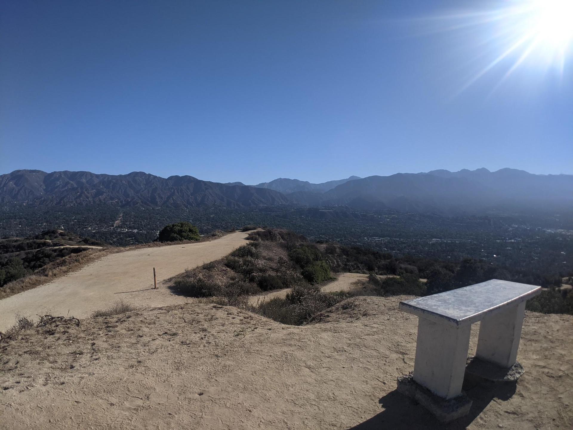 Sunny mountain view with a dirt path and a stone bench overlooking a valley.