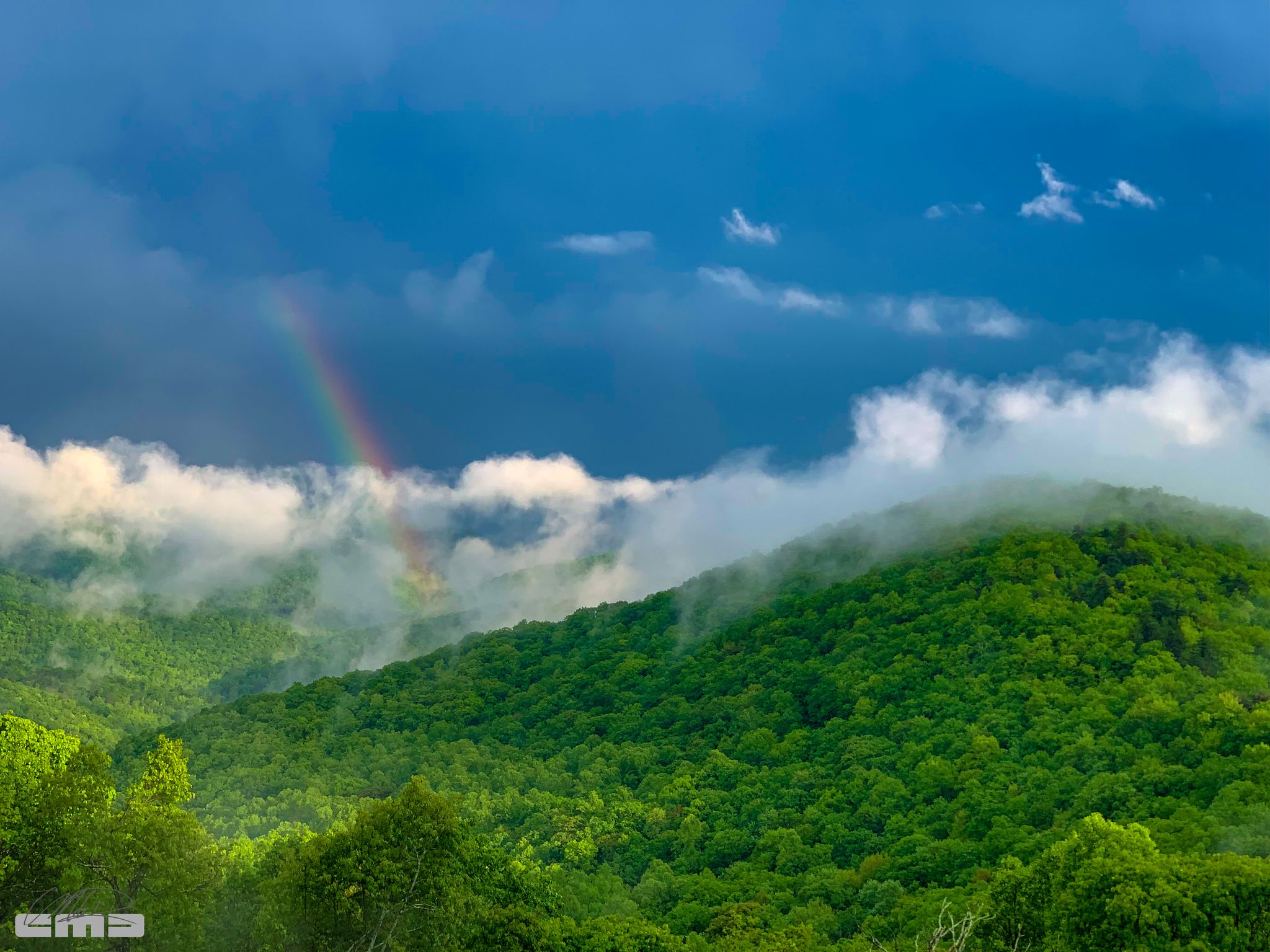 A rainbow arches over misty, forested mountains under a dramatic sky. - Mountains to Sea Bicycle Route