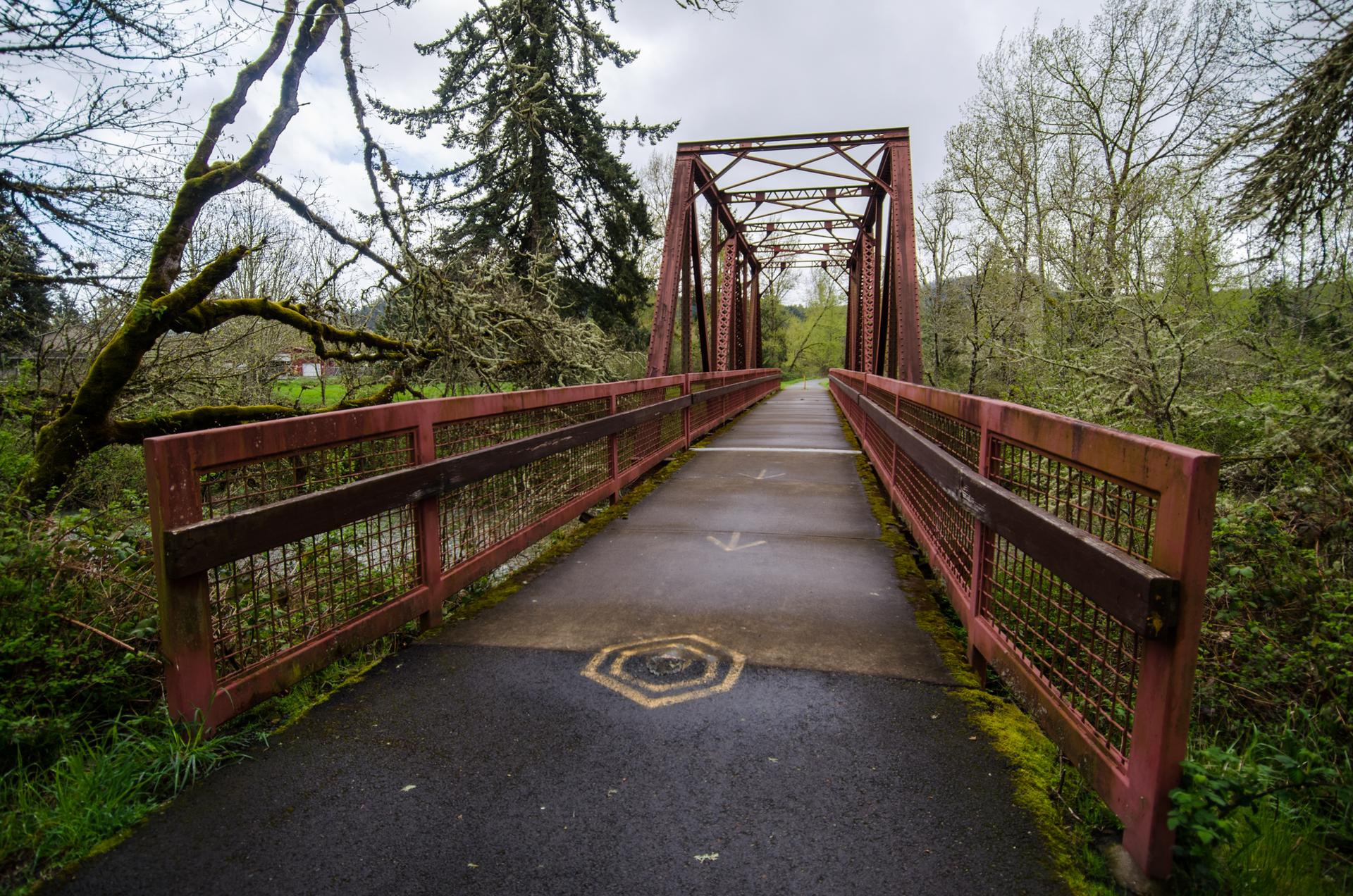 Red metal truss bridge over a path, surrounded by green trees.