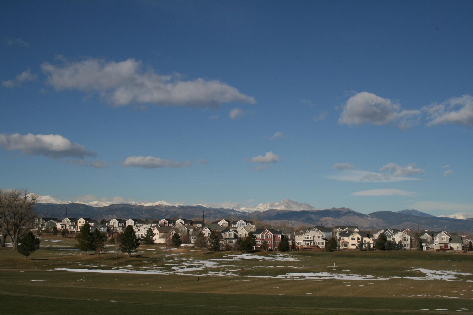 Suburban houses under a blue sky, backed by snow-capped mountains and green fields.