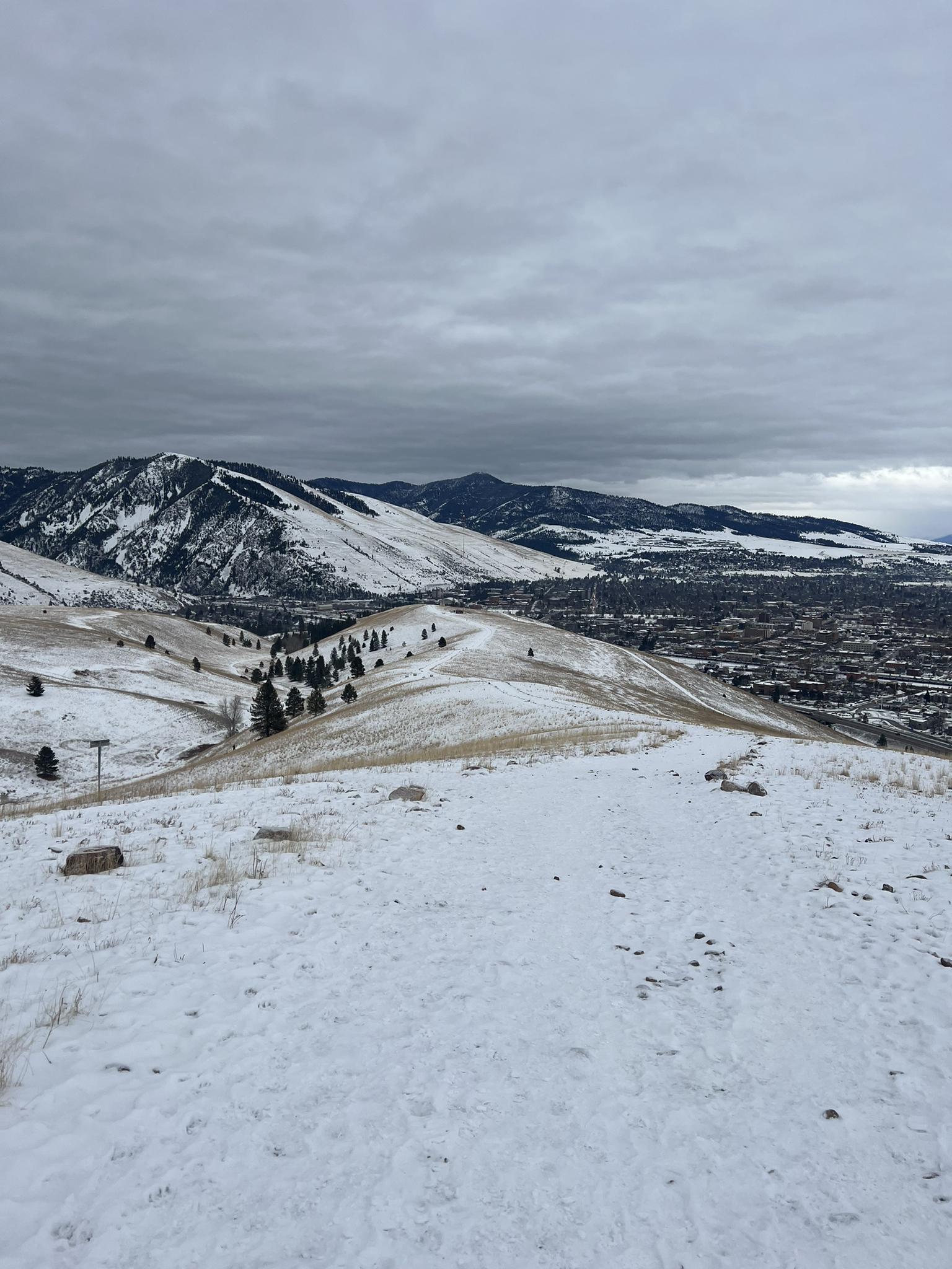 Snowy hills and mountains overlook a distant town under an overcast sky.
