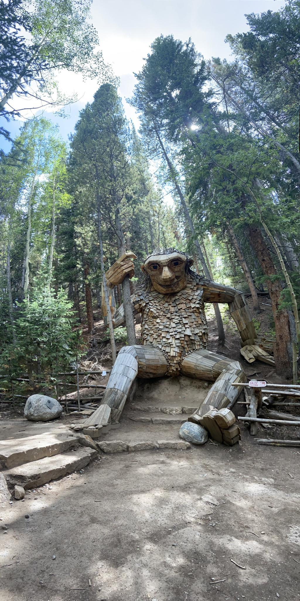 A large wooden troll sculpture sits in a forest clearing on a bright day.