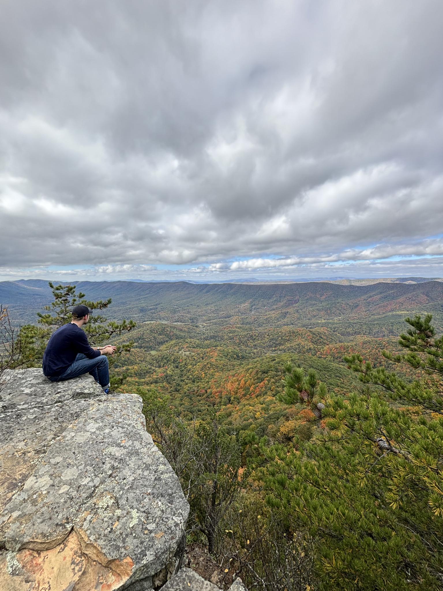 Man sits on a cliff overlooking vast, cloudy, autumn-colored mountains.
