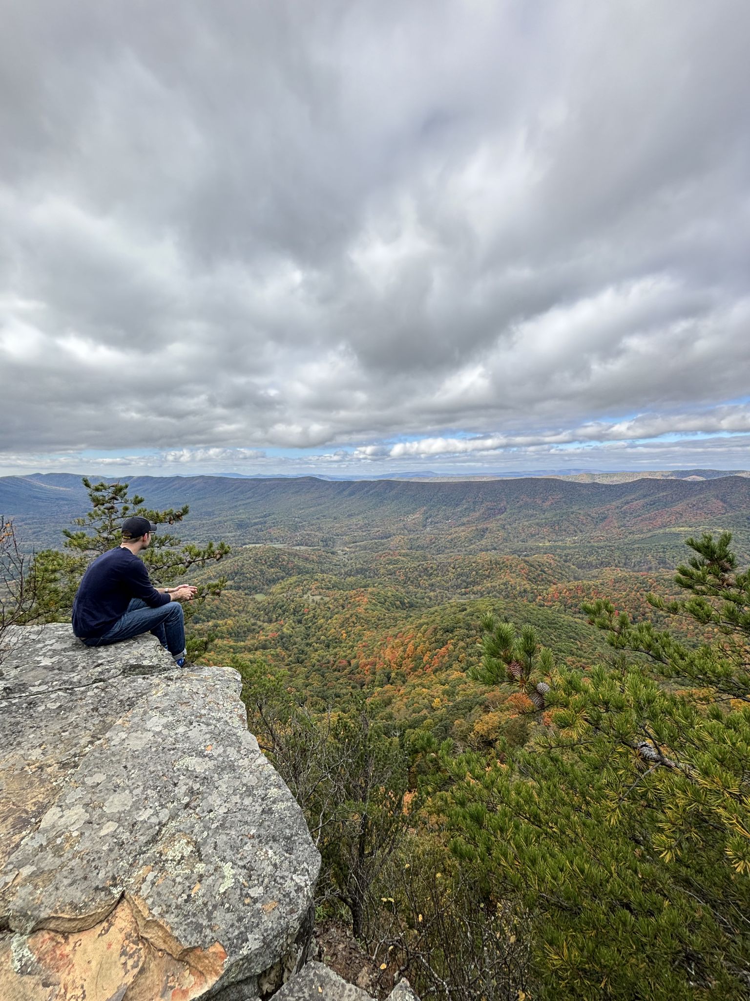 Man sits on a cliff overlooking vast, cloudy, autumn-colored mountains. - Big House Mountain Trail