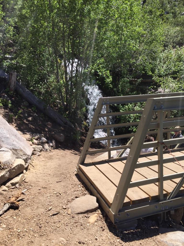 A small wooden and metal bridge crosses a flowing waterfall in a green forest.