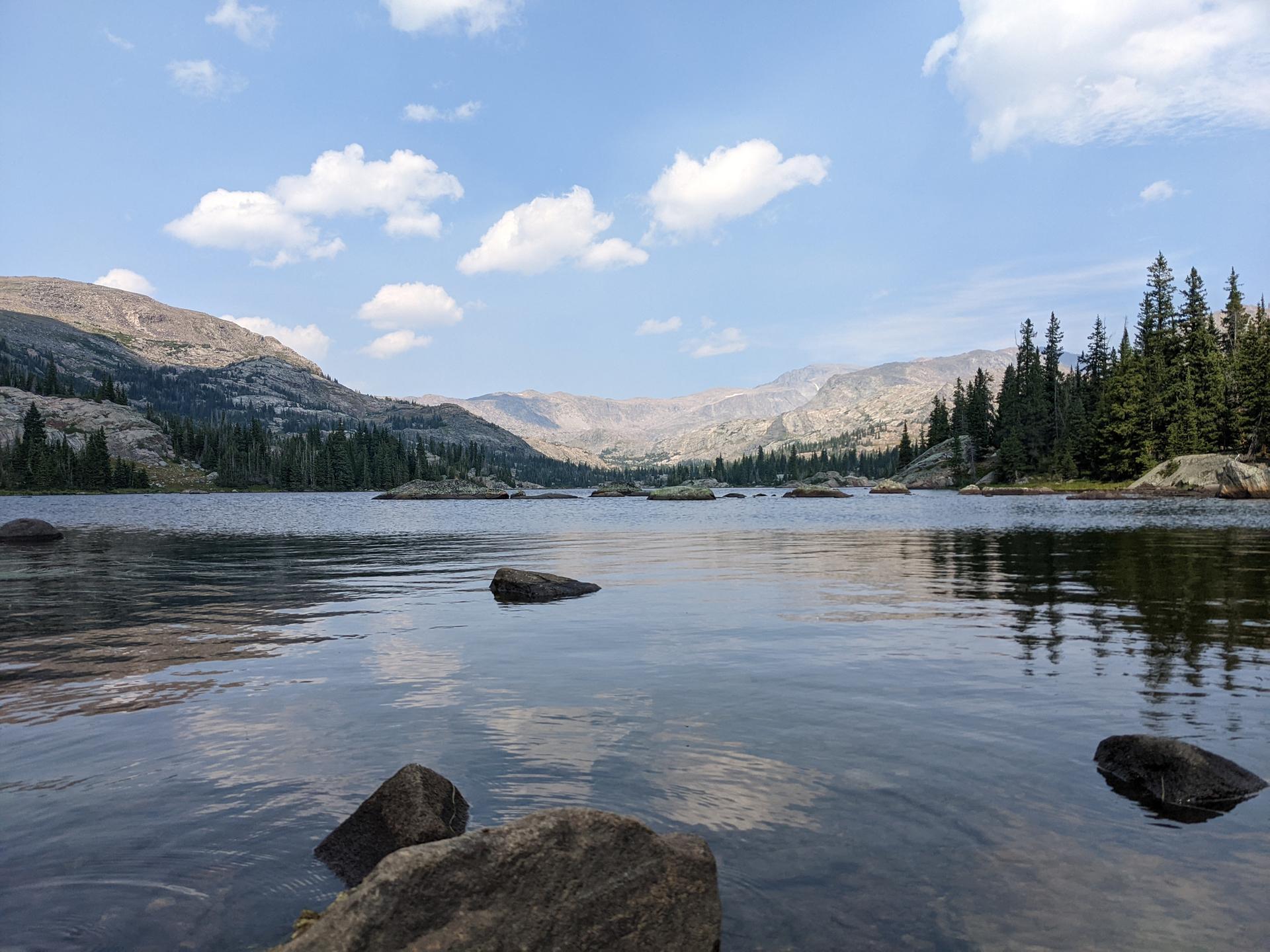 Wide lake surrounded by mountains and pine forests under a cloudy sky.